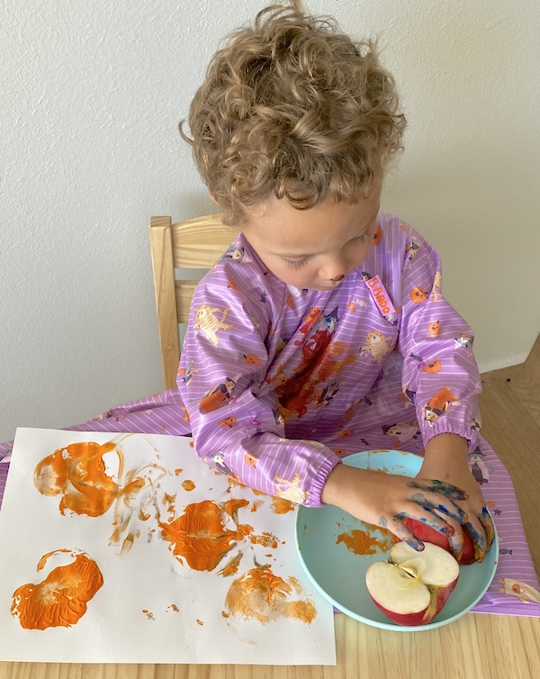 Boy using an apple to stamp orange paint on paper. 