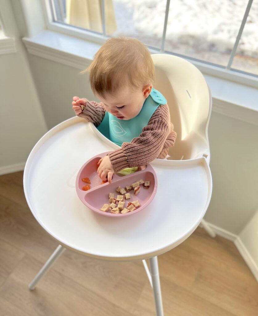 Baby girl self feeding food in high chair from a pink plate